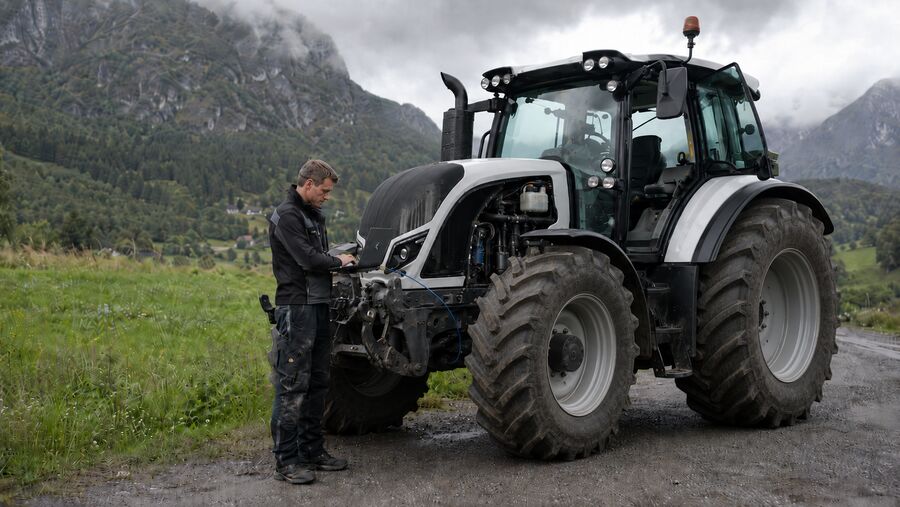 Teknikar brukar diagnoselaptop ved ein moderne traktor på ein rural veg med fjell i bakgrunnen